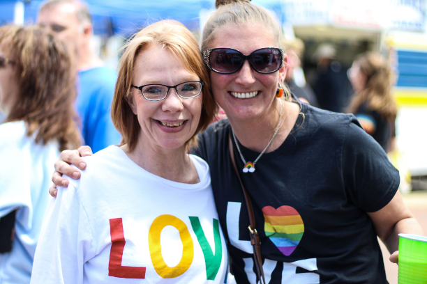 Two women smiling and embracing at an outdoor festival, celebrating love and friendship under the sun.