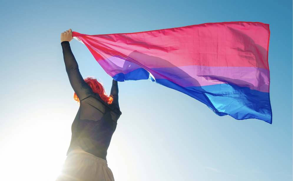 A red haired person holds up a bisexual flag that waves in the wind.