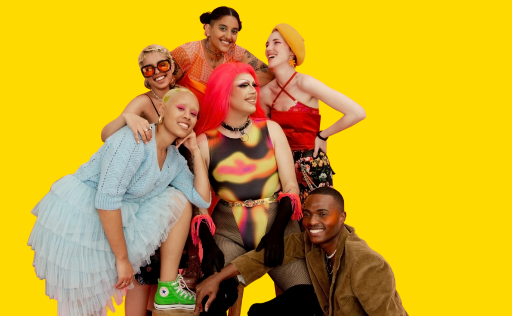 A group of young, diverse queer people laugh and smile in front of a yellow background.